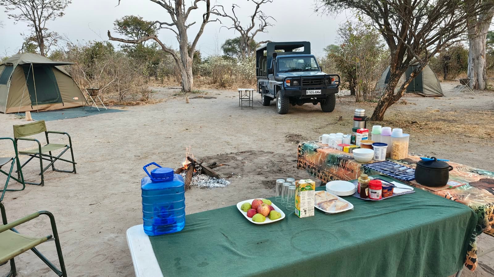 Mobile safari camp breakfast table with campfire, tents, and safari vehicle in the Botswana bush