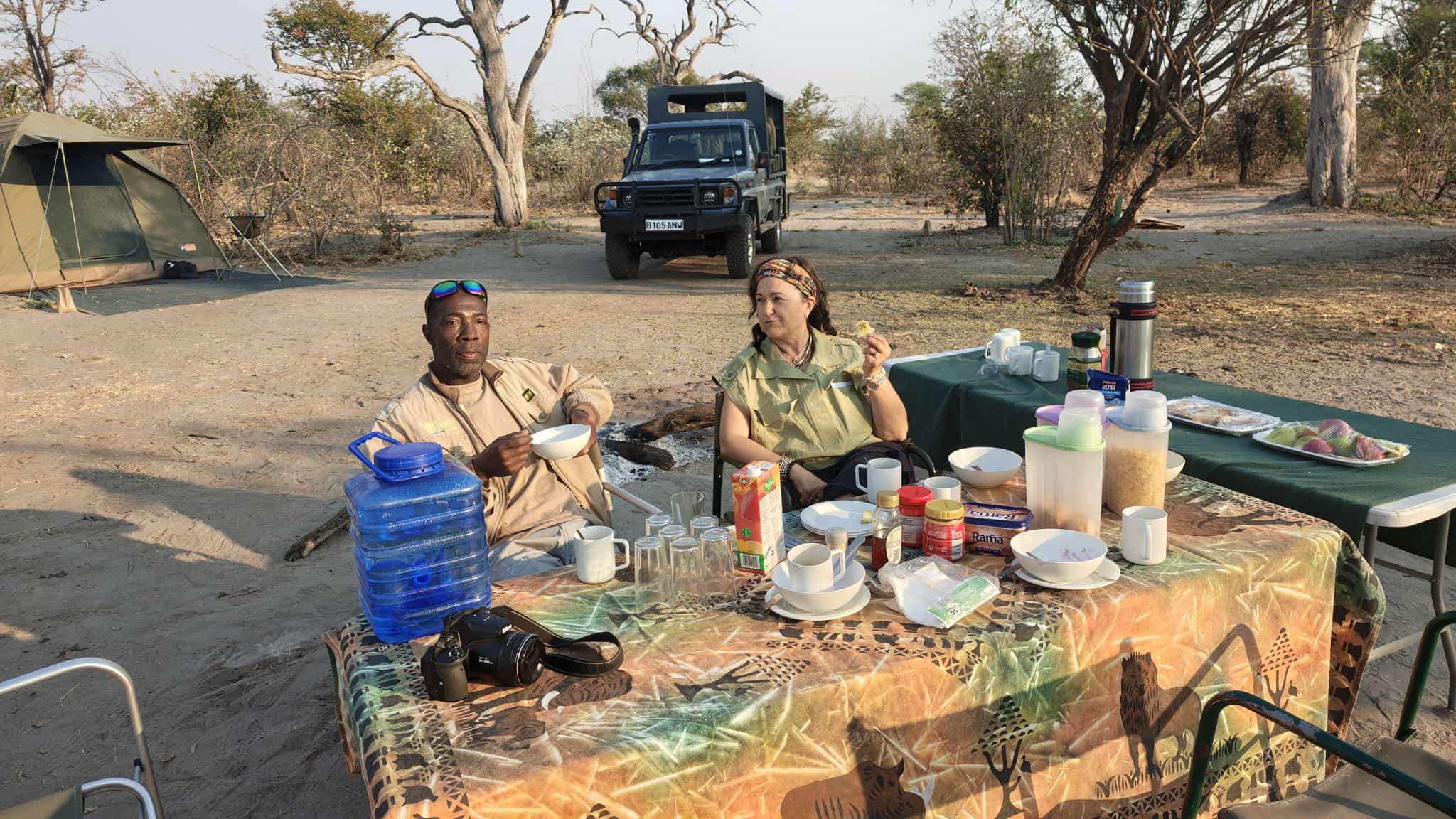Guide and guest sharing a meal at the camp table with safari vehicle in background